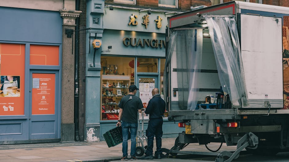 Two movers from West Kensington Removals are engaged in the loading process outside a shopfront named 'Guangh' on a city street. One mover, dressed in dark clothing, is holding a dolly and is position