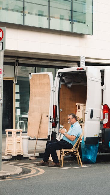 A man wearing glasses and a light blue shirt sits on a wooden chair outside a white moving van parked on a city street. The van's rear door is open, revealing cardboard boxes, wooden furniture panels,