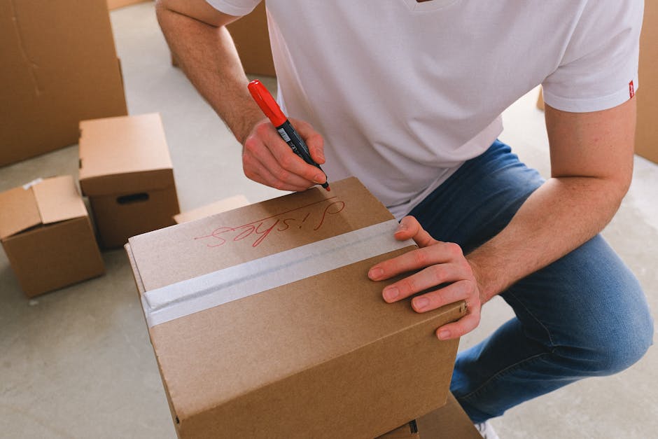 A person wearing a white t-shirt and blue jeans is kneeling inside a house, using a red marker to label a small cardboard box with the word 'shelves' written in red ink. The box is sealed with white p