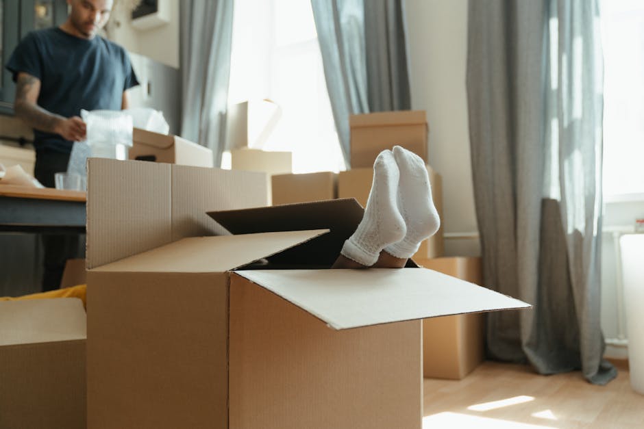 A close-up view of a pair of feet wearing white socks protruding from inside an open cardboard moving box, which is part of a home relocation process. The box is situated on a light wooden floor in a 