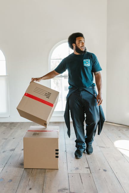 Two movers from West Kensington Removals are engaged in a home relocation process inside a well-lit room with white walls and wooden flooring. The first mover, positioned on the right, is carrying a l