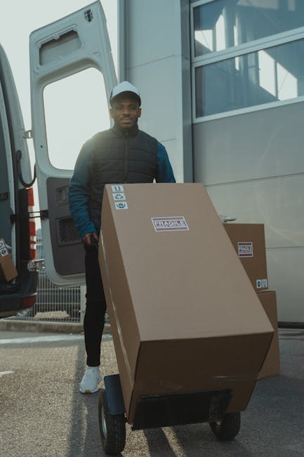 A man wearing a black jacket, blue sleeves, a light blue cap, and white sneakers is standing outdoors next to a large white van with an open rear door. He is positioned beside a black hand truck loade