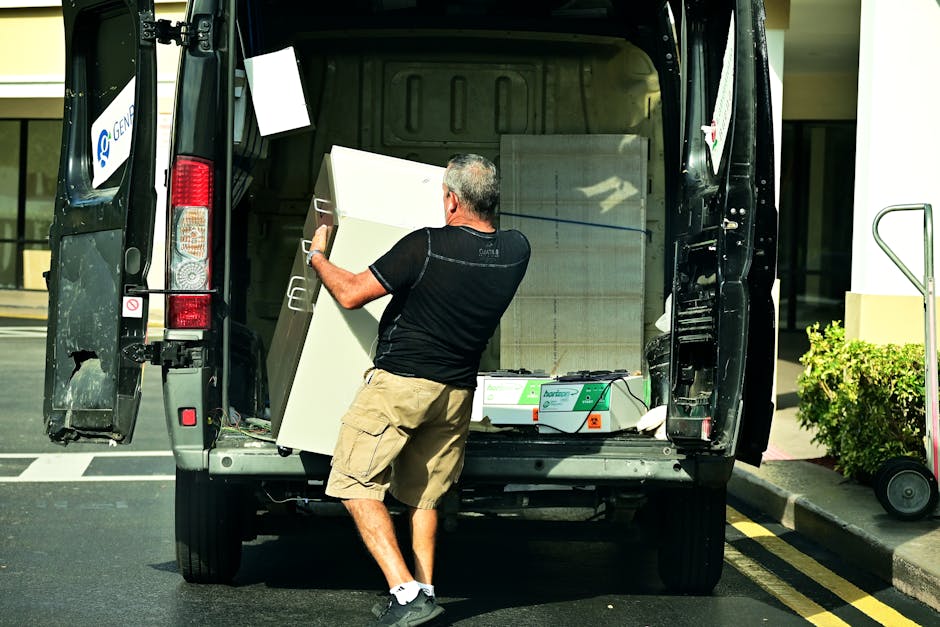 A man wearing a black jacket, blue sleeves, a light blue cap, and white sneakers is standing outdoors next to a large white van with an open rear door. He is positioned beside a black hand truck loade